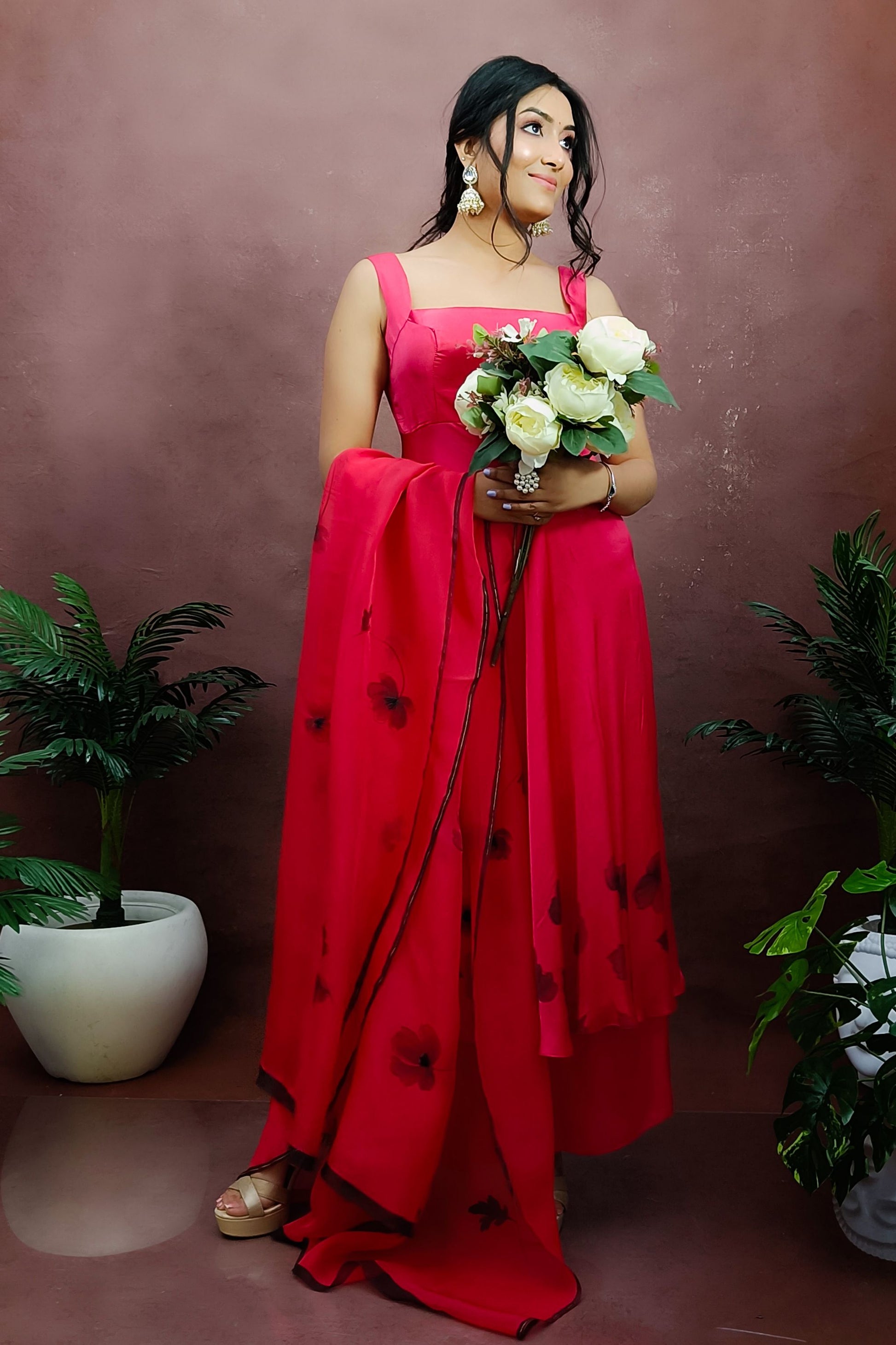 Girl wearing a red colour sleeveless anarkali suit enhanced with hand painted black poppy flowers adorned with delicate bead work., paired with organza handpainted dupatta and a flared palazzo.