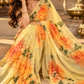 A woman wearing a yellow floral saree with orange and green flower prints, sitting on stone steps in an outdoor setting.