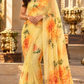 A woman wearing a yellow saree with orange and green floral patterns, standing in front of an ornate architectural background with gold-colored decorative elements.