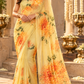 A woman models a yellow saree with a vibrant orange and green floral print in an outdoor architectural setting with stone steps and gold decorative urns.