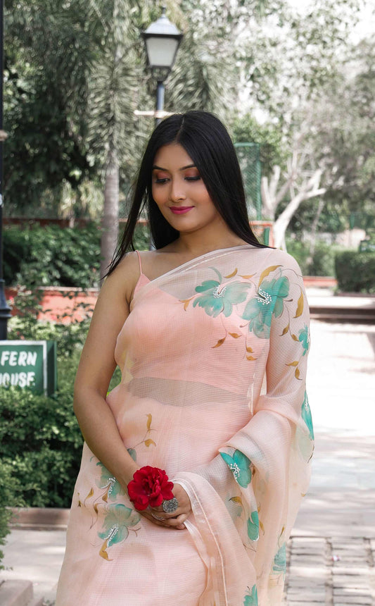 Graceful hand-painted Kota Doria Silk Saree in light peach, adorned with green floral motifs, and beautifully enhanced with sequin and pearl hand embroidery. A true masterpiece of hand painting on saree, showcasing the timeless beauty of pure Kota silk